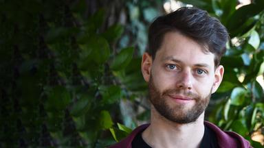 Ein junger Mann mit Bart und braunem Haar steht vor einem Hintergrund aus grünen Pflanzen und blickt mit lächelnd in die Kamera.