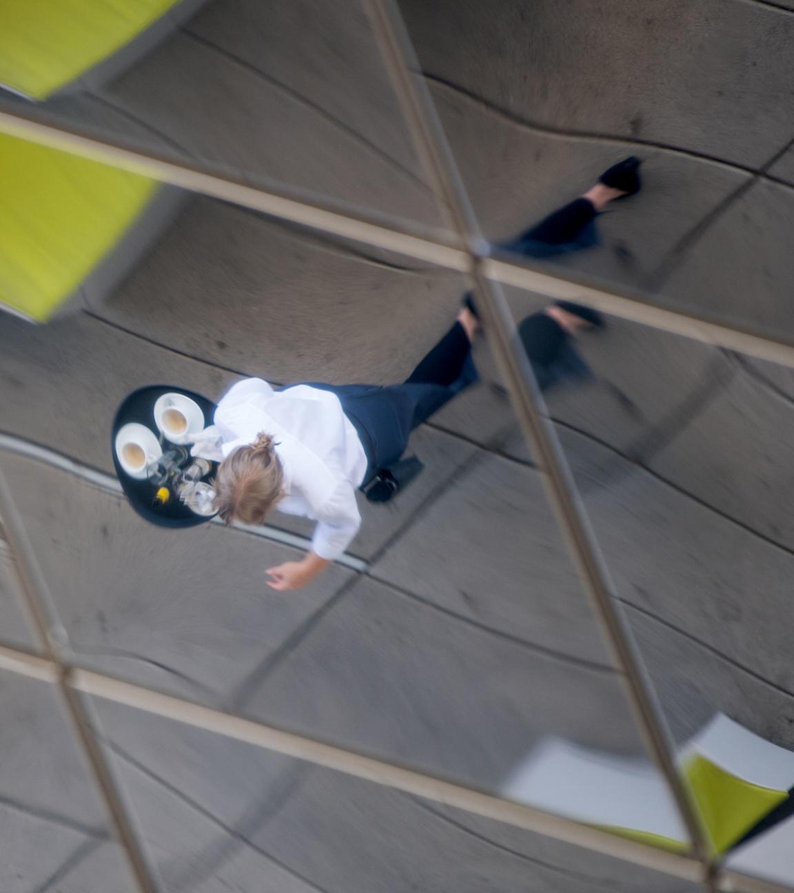 Archiv: Eine Frau, die Kaffee auf einem Tablett trägt, spiegelt sich am 13.07.2017 im einer verspiegelten Außendecke des Porsche-Museums in Stuttgart.
