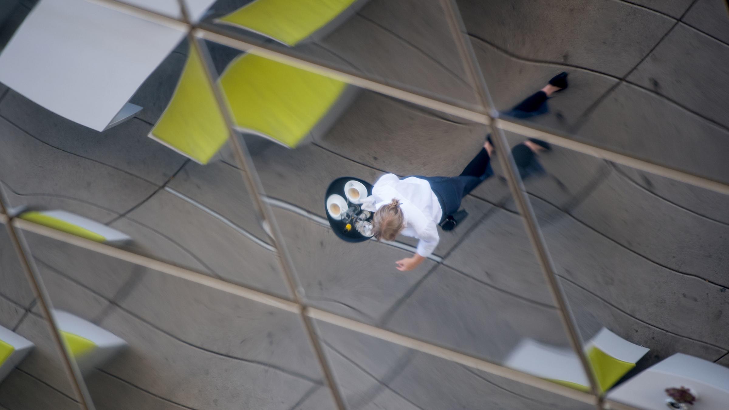 Archiv: Eine Frau, die Kaffee auf einem Tablett trägt, spiegelt sich am 13.07.2017 im einer verspiegelten Außendecke des Porsche-Museums in Stuttgart.