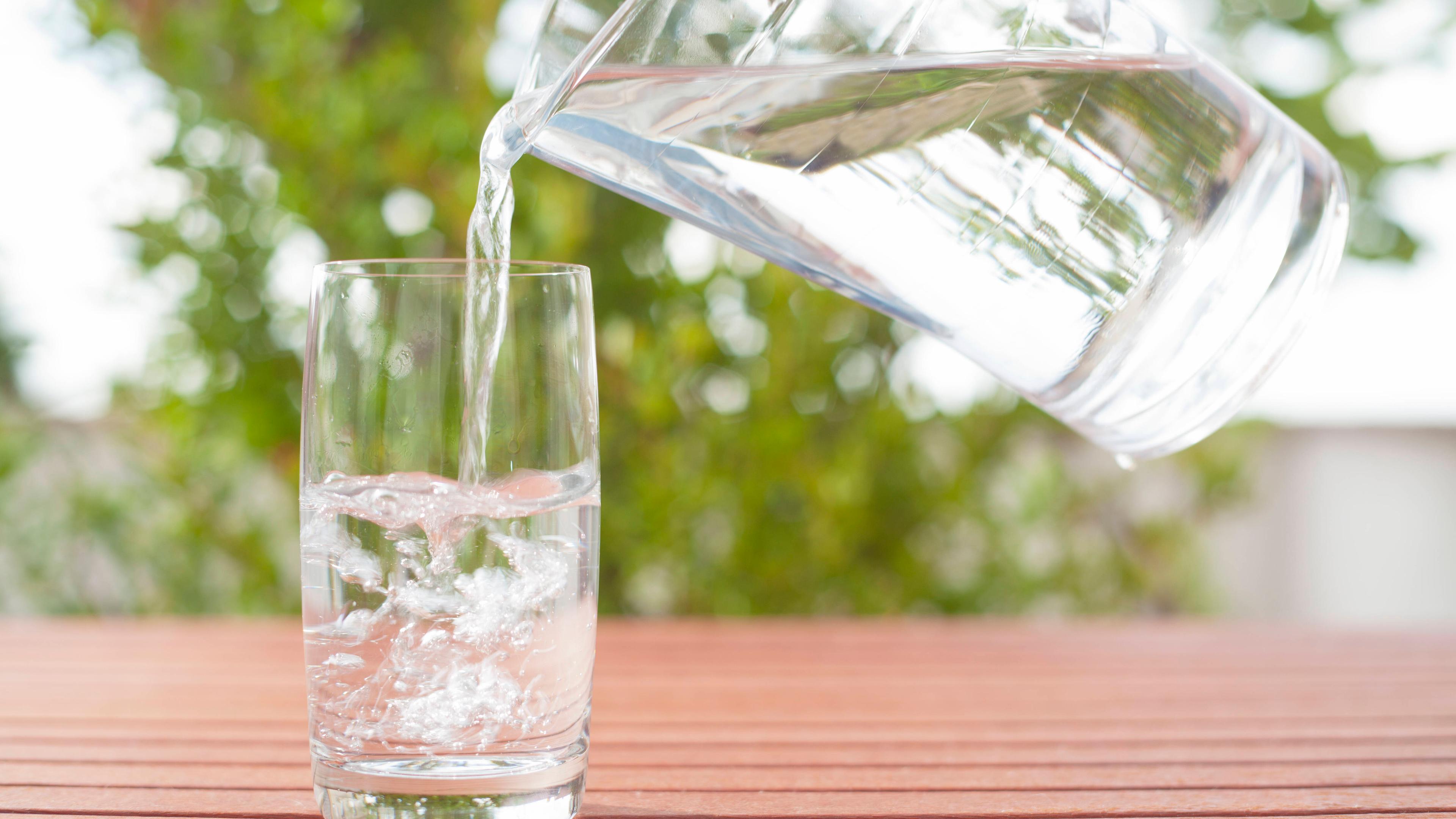 Eine Person schenkt stilles Wasser aus einem Krug in ein Glas ein, das auf einem Gartentisch steht.