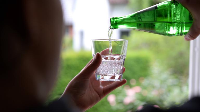 Eine Frau steht am Fenster und gießt Mineralwasser aus einer Flasche in ein Glas.