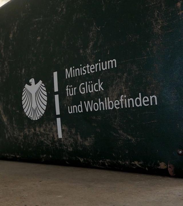 Koffer mit der Aufschrift "Ministerium für Glück und Wohlbefinden"