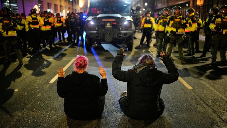 Zwei Personen sitzen während einer Protestaktion mit erhobenen Händen vor der Minnesota State Patrol auf der Straße.