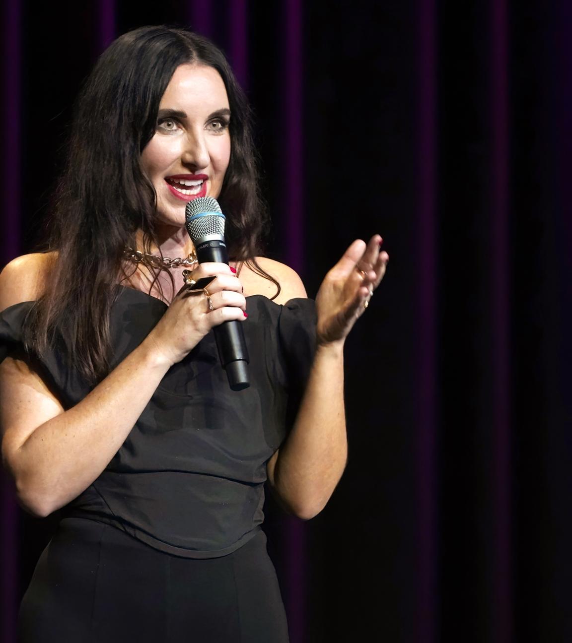 Eine junge Frau mit langen dunklen Haaren steht auf einer dunklen Bühne und spricht in ein Mikrofon.