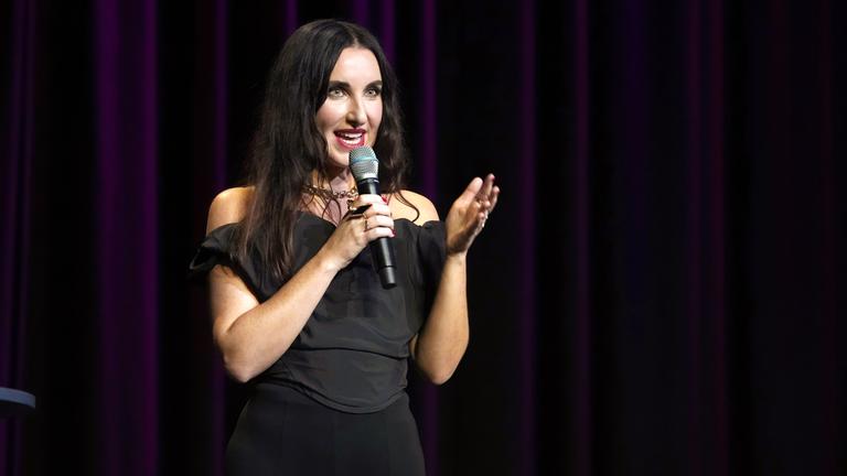 Eine junge Frau mit langen dunklen Haaren steht auf einer dunklen Bühne und spricht in ein Mikrofon.