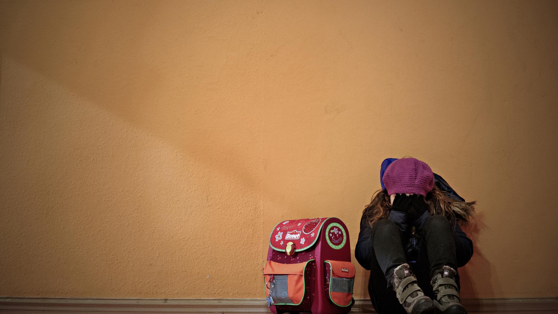 Ein Mädchen sitzt neben ihrem Schulranzen auf dem Boden und hält die Hände vor das Gesicht.