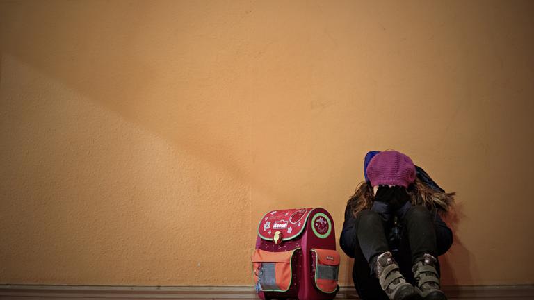 Ein Mädchen sitzt neben ihrem Schulranzen auf dem Boden und hält die Hände vor das Gesicht.