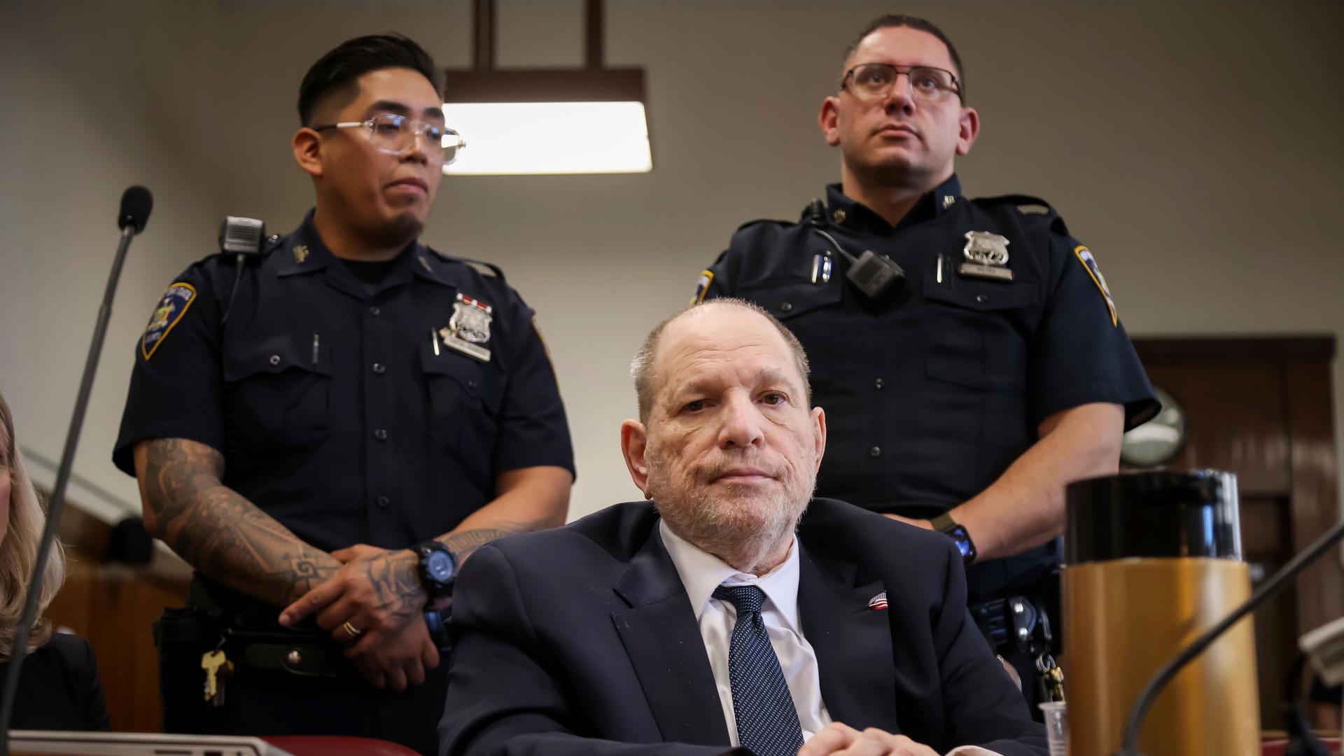 Harvey Weinstein sitzt in einem Gerichtssaal, hinter ihm stehen zwei Polizisten.