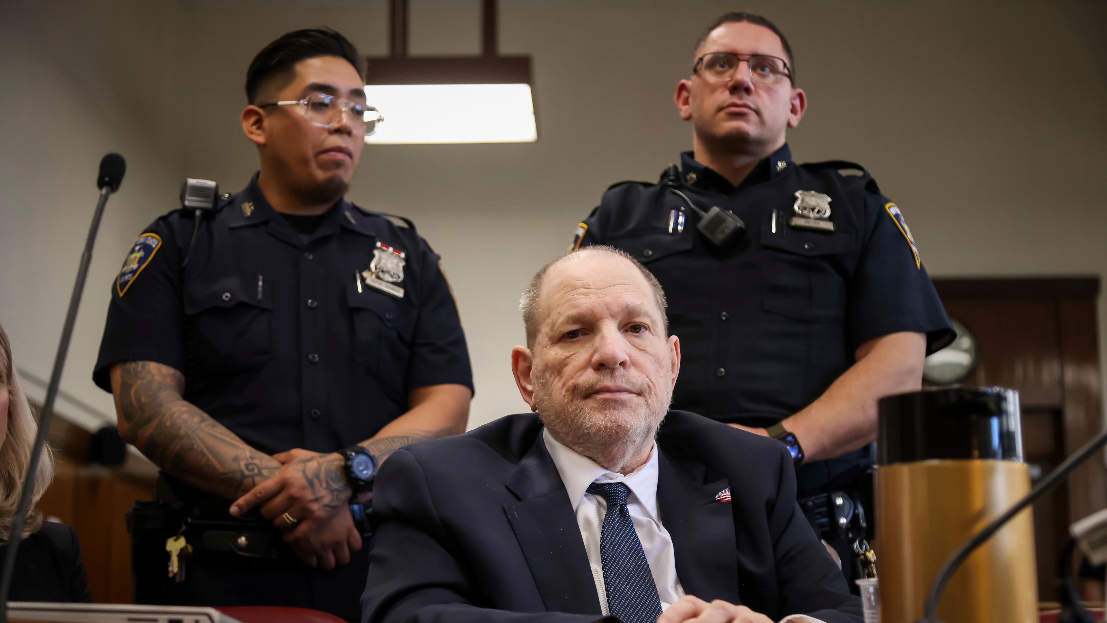 Harvey Weinstein sitzt in einem Gerichtssaal, hinter ihm stehen zwei Polizisten.
