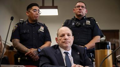 Harvey Weinstein sitzt in einem Gerichtssaal, hinter ihm stehen zwei Polizisten.