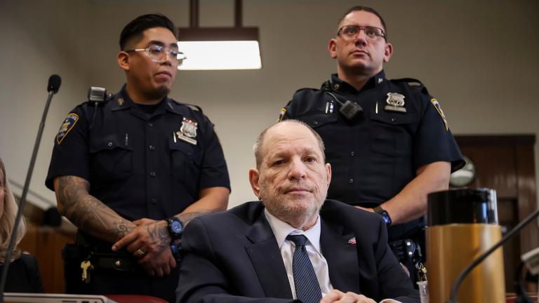 Harvey Weinstein sitzt in einem Gerichtssaal, hinter ihm stehen zwei Polizisten.