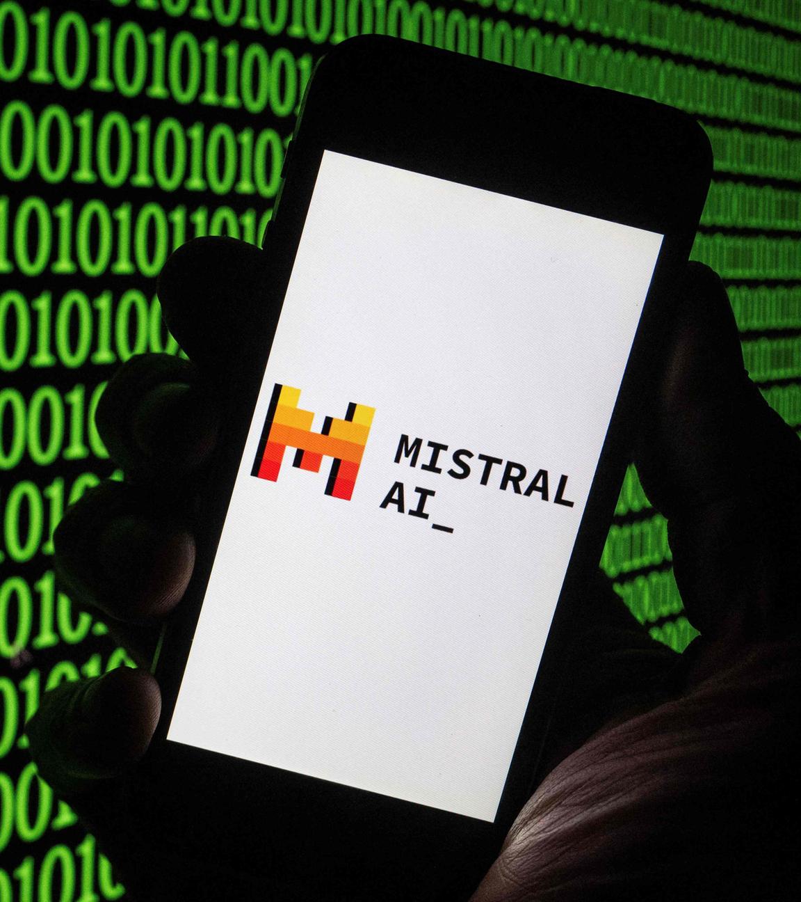 Eine Hand hält ein Smartphone auf dem das Logo des französischen Start-ups Mistral AI mit einem rot-gelben M zu sehen ist. Im Hintergund leuchtet grüner Binärcode.