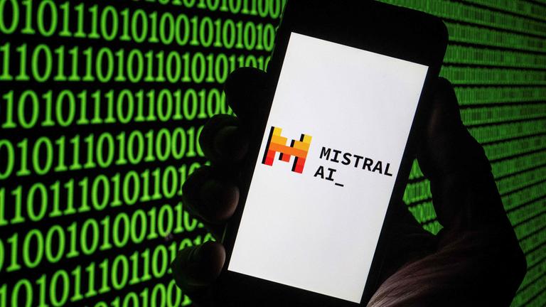 Eine Hand hält ein Smartphone auf dem das Logo des französischen Start-ups Mistral AI mit einem rot-gelben M zu sehen ist. Im Hintergund leuchtet grüner Binärcode.