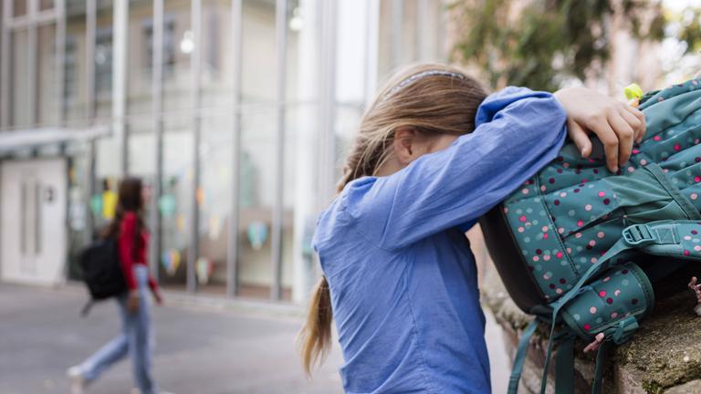 Ein Mädchen verdeckt ihr Gesicht in ihrem Ranzen während ein anderes Mädchen an ihr vorbeiläuft.