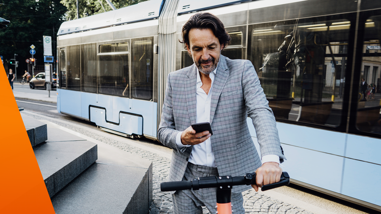Mann im Anzug schaut ins Handy und steht bei einem E-Roller