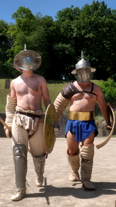Zwei Männer mit Schild, Schwert und Helm in Gladiatoren-Uniform laufen bei Sonnenschein durch eine sandige Arena. 