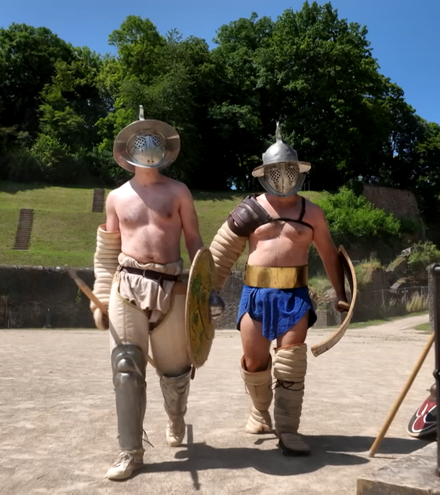 Zwei Männer mit Schild, Schwert und Helm in Gladiatoren-Uniform laufen bei Sonnenschein durch eine sandige Arena. 