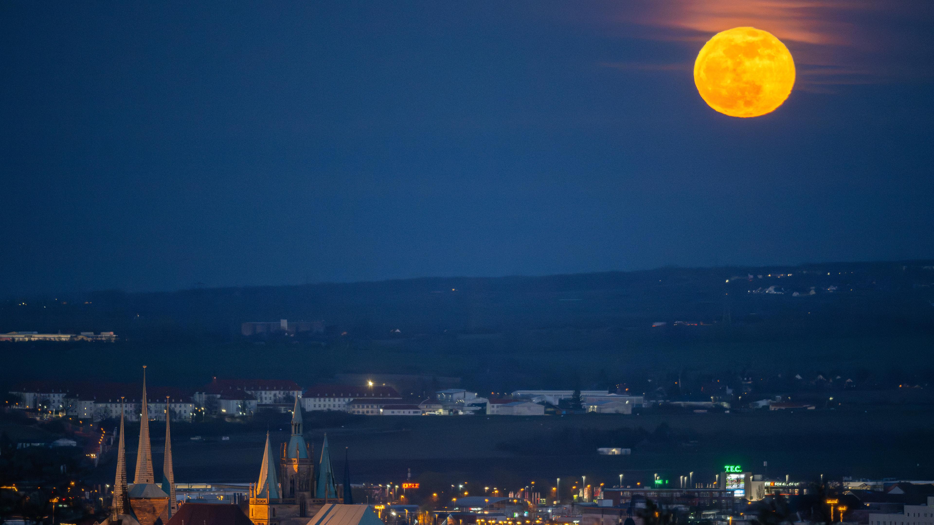 Der rötlich, gelb strahlende Vollmond geht am Abend am Horizont über der Landeshauptstadt Erfurt auf am 25.03.2024