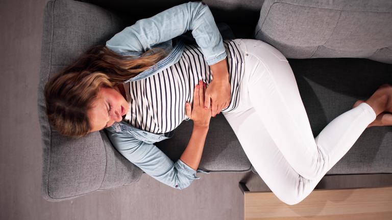 Eine Frau liegt auf dem Sofa und hält sich ihren Bauch mit einem von Schmerzen verzerrten Gesicht.
