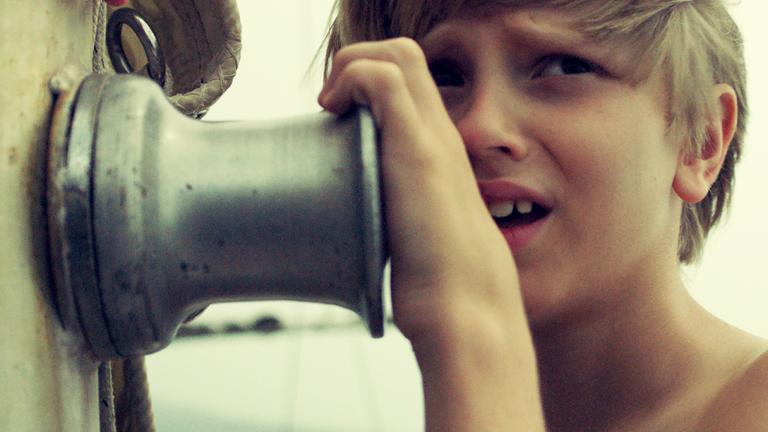Spielszene: Ein Junge mit panischem Blick umklammert eine Winde auf einem Segelboot.