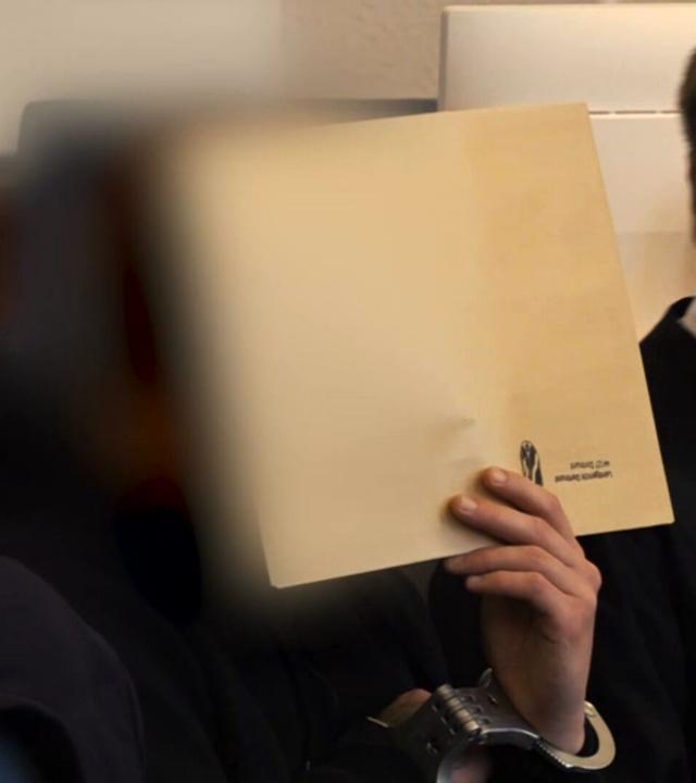 Ein Mann sitzt im Gerichtssaal und versteckt sein Gesicht hinter einer Mappe.