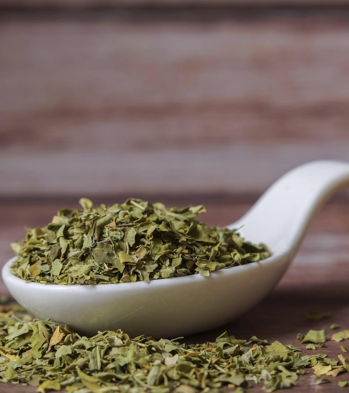 Getrocknete Moringa Oleifera Blätter liegen in einem weißen Porzellanlöffel auf einem Holztisch.