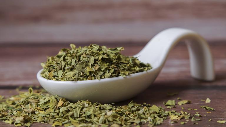 Getrocknete Moringa Oleifera Blätter liegen in einem weißen Porzellanlöffel auf einem Holztisch.