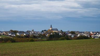 Blick auf ein kleines Dorf mit Kirche hinter einem Feld, auf dem es grün sprießt. Darüber blauer Himmel.