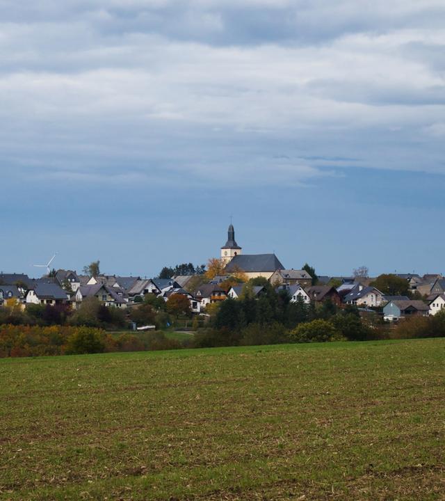 Blick auf ein kleines Dorf mit Kirche hinter einem Feld, auf dem es grün sprießt. Darüber blauer Himmel.