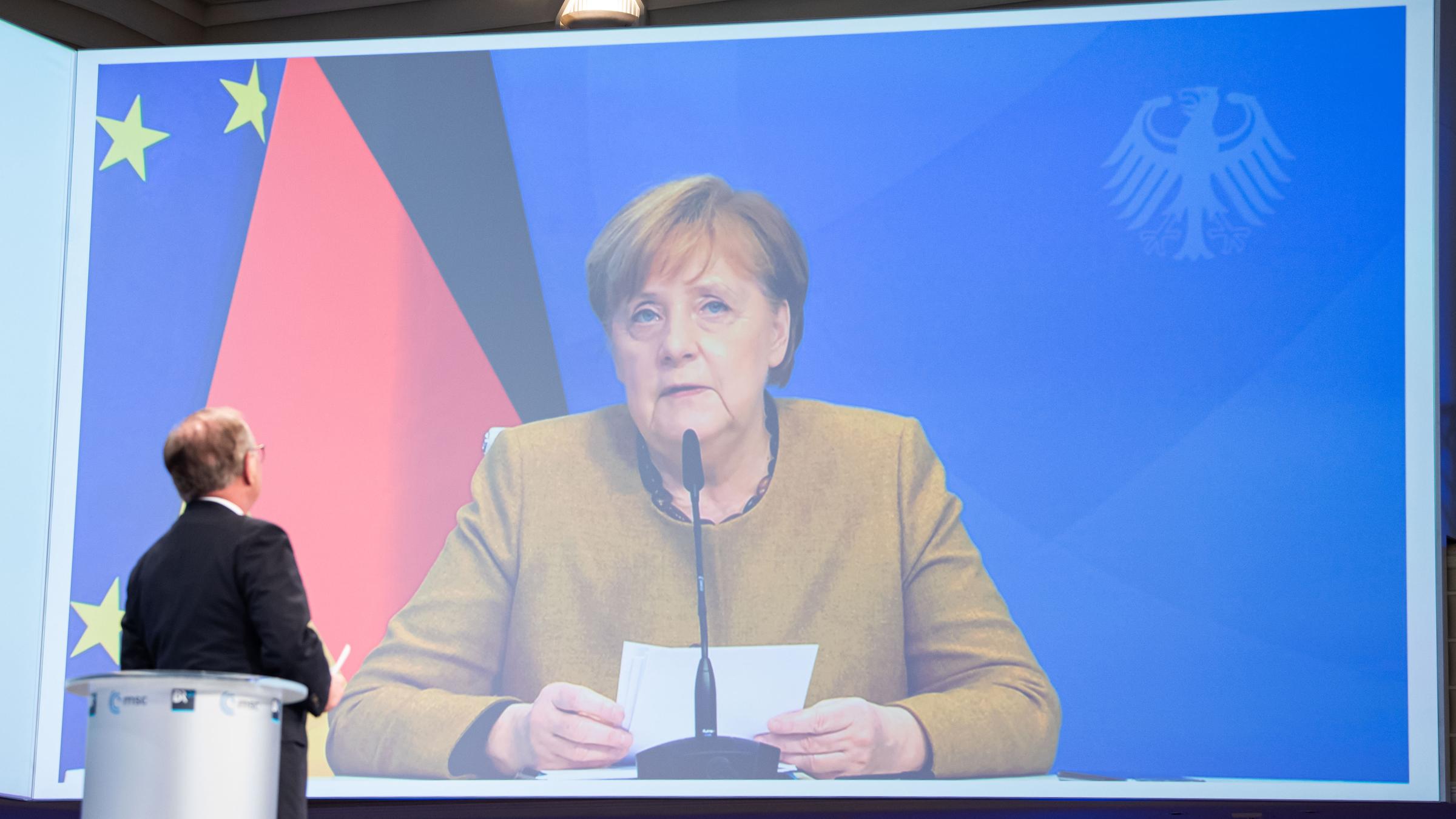 Zu sehen ist Angela Merkel auf einer großen Leinwand, wie sie sitzend eine Rede hält. Hinter ihr sieht man die deutsche Flagge.