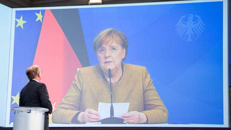 Zu sehen ist Angela Merkel auf einer großen Leinwand, wie sie sitzend eine Rede hält. Hinter ihr sieht man die deutsche Flagge.
