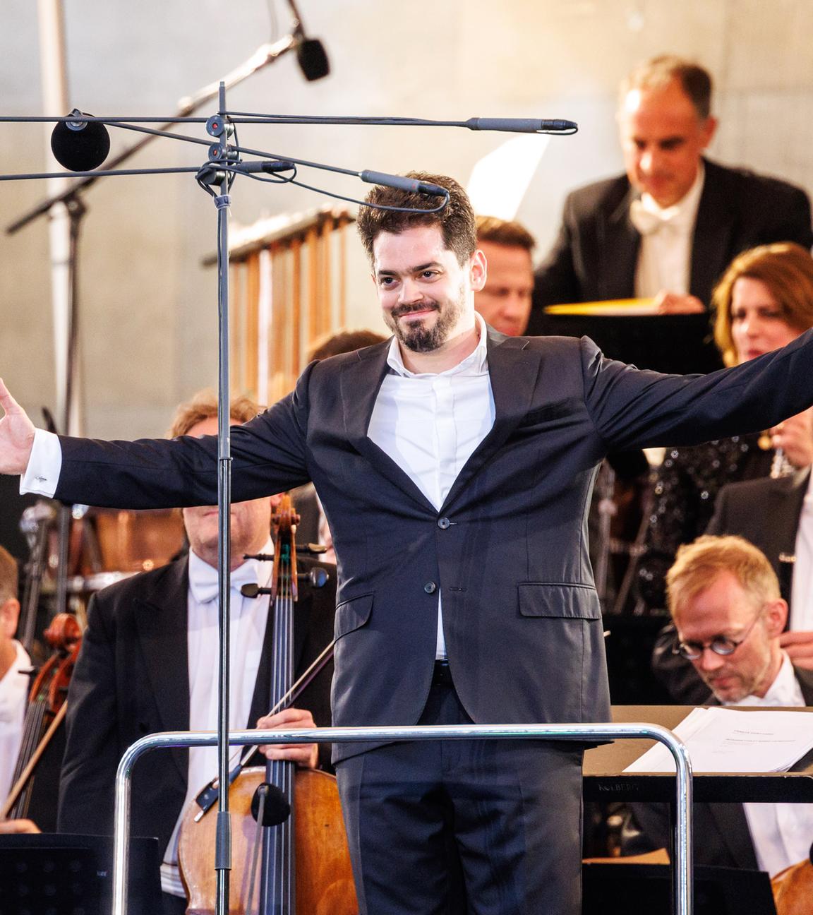 Ein Dirigent steht mit ausgebreiteten Armen vor seinem Orchester und lächelt dem Publikum zu.