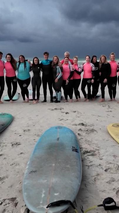 Auf dem Bild sind mehere Frauen zu sehen am Strand mit ihrer Surfausrüstung.