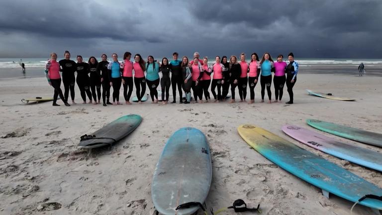 Auf dem Bild sind mehere Frauen zu sehen am Strand mit ihrer Surfausrüstung.