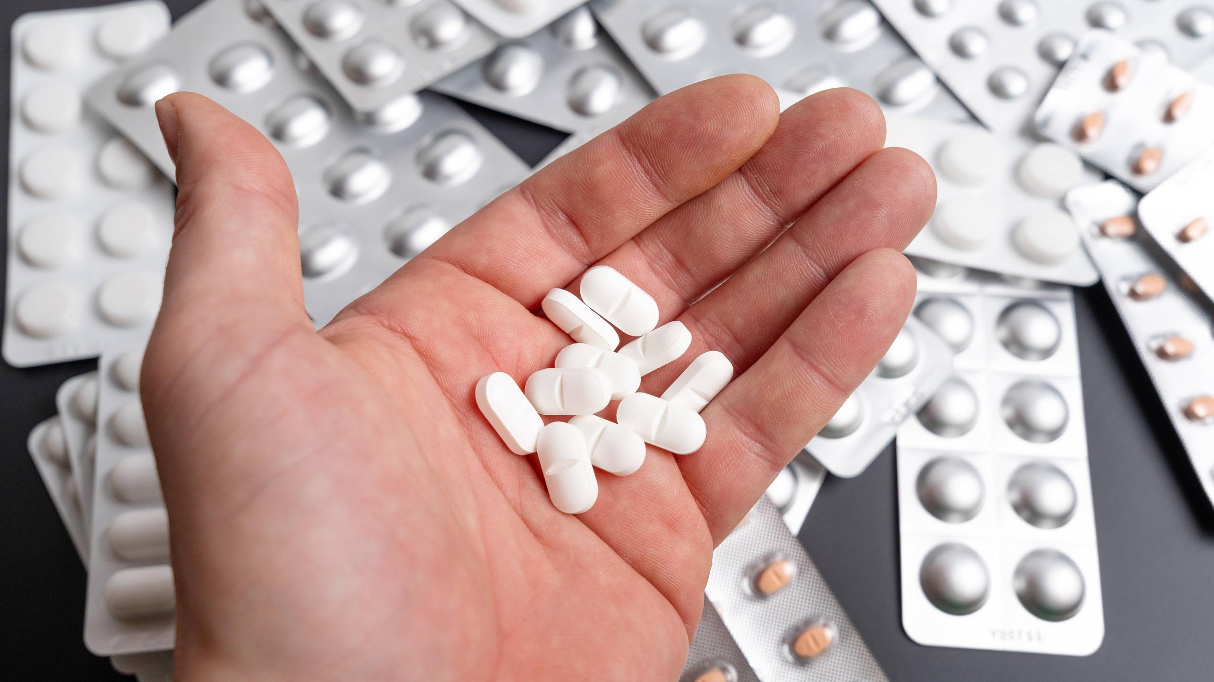 Eine Hand hält viele Tabletten, im Hintergrund sind Blister zu sehen.