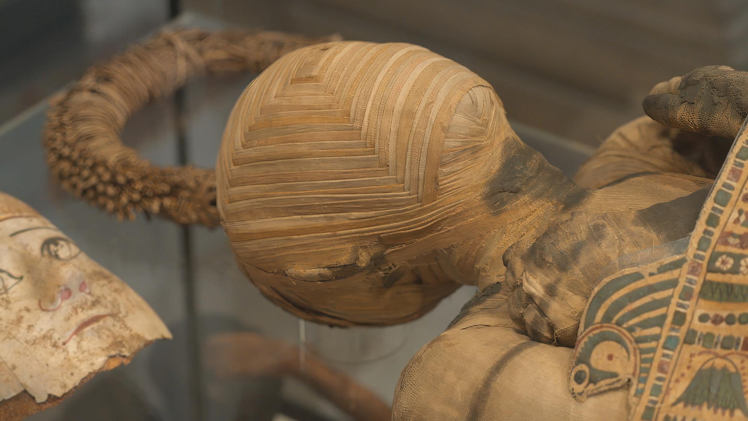 Eine ägyptische Mumie mit bandagiertem Kopf liegt auf einem Glastisch, auf der Brust liegt eine bemalte Kartonage.