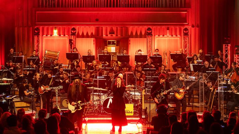 Man sieht einen beleuchteten Konzertsaal mit einer Band, einer Sängerin, einem Orchester und Publikum.