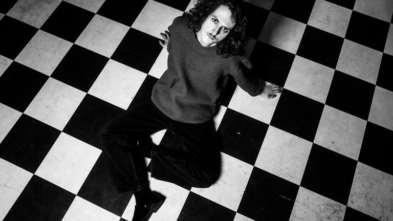 Auf einer schwarz-weiß-Fotografie sieht man einen jungen Mann der auf einem schwarz-weiß-gefliestem Boden sitzt.