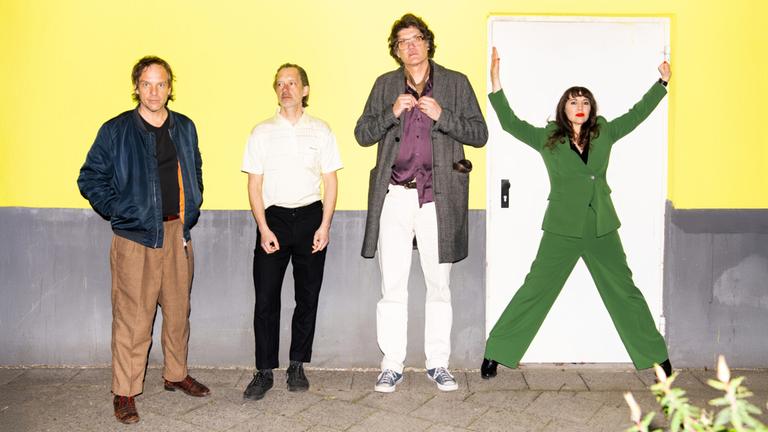 Drei Männer und eine Frau stehen vor einer gelb-grauen Wand.