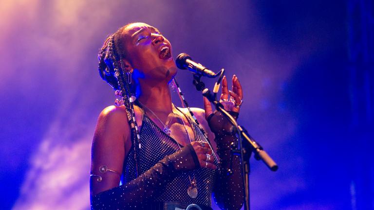 Eine schwarze Frau in glitzerndem Kostüm singt auf einer Bühne.