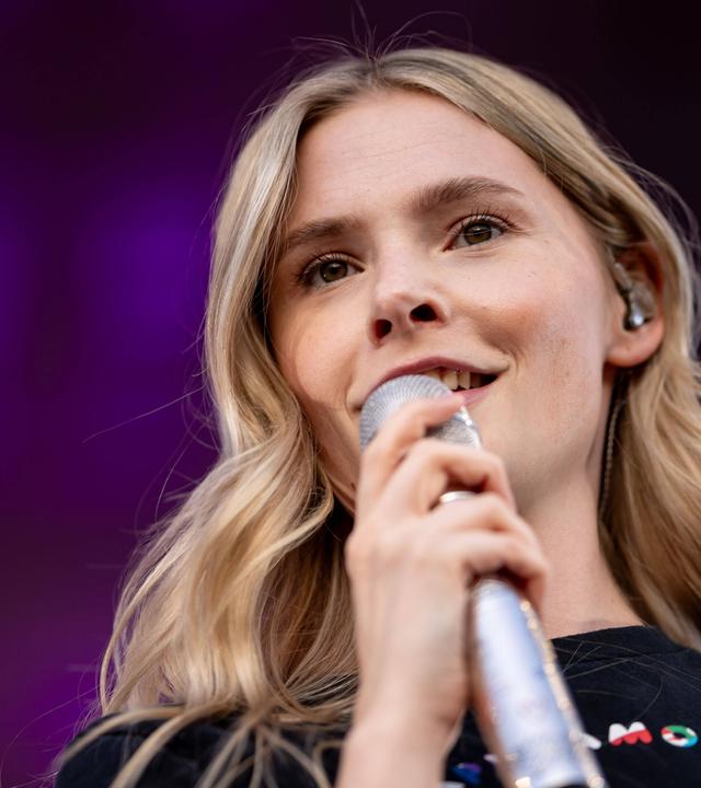 Porträtfoto einer jungen blonden Frau mit langen Haaren, die in ein Mikrofon singt.