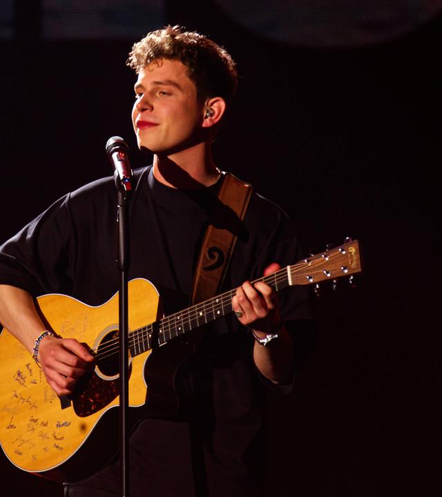 Ein junger Mann spielt Gitarre auf einer Bühne und singt in ein Mikrofon.