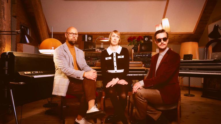 Zwei Männer und eine Frau sitzen in einem Musikstudio im Stil der 1970er Jahre.