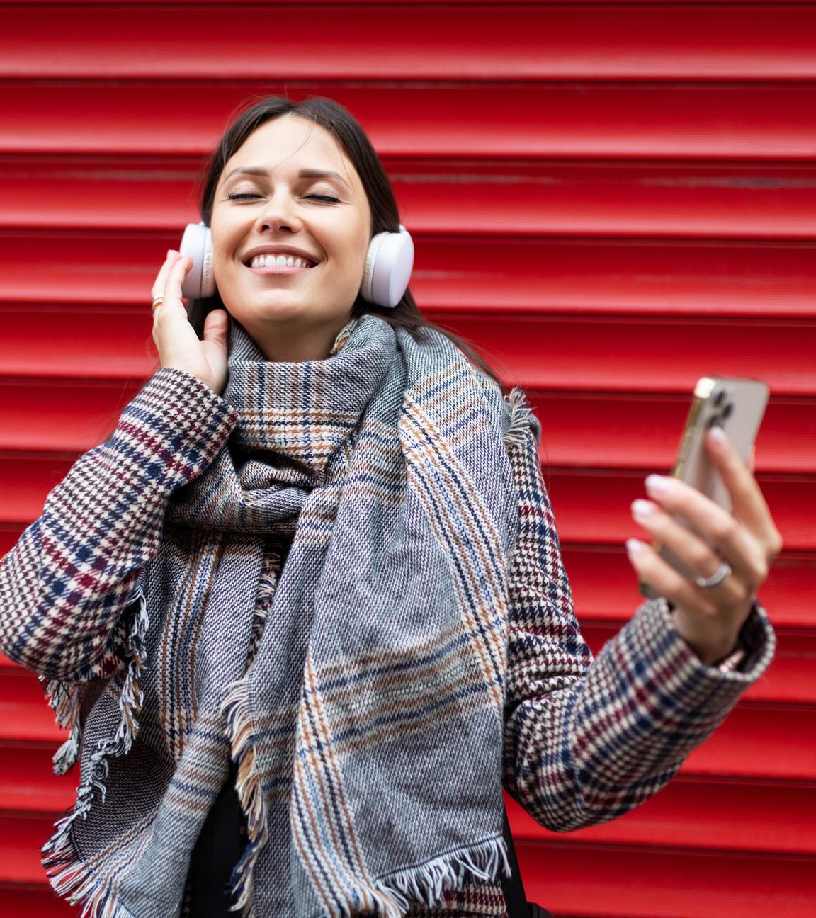 Junge Frau hört vor einer roten Wand Musik mit ihren Kopfhörern sie hält ein Smartphone in der Hand