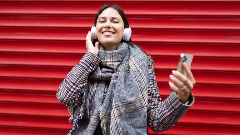 Junge Frau hört vor einer roten Wand Musik mit ihren Kopfhörern sie hält ein Smartphone in der Hand