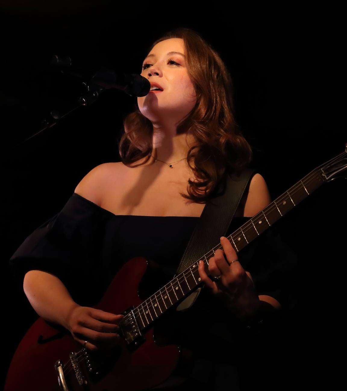Eine Frau steht auf einer Bühne, spielt Gitarre und singt dazu.
