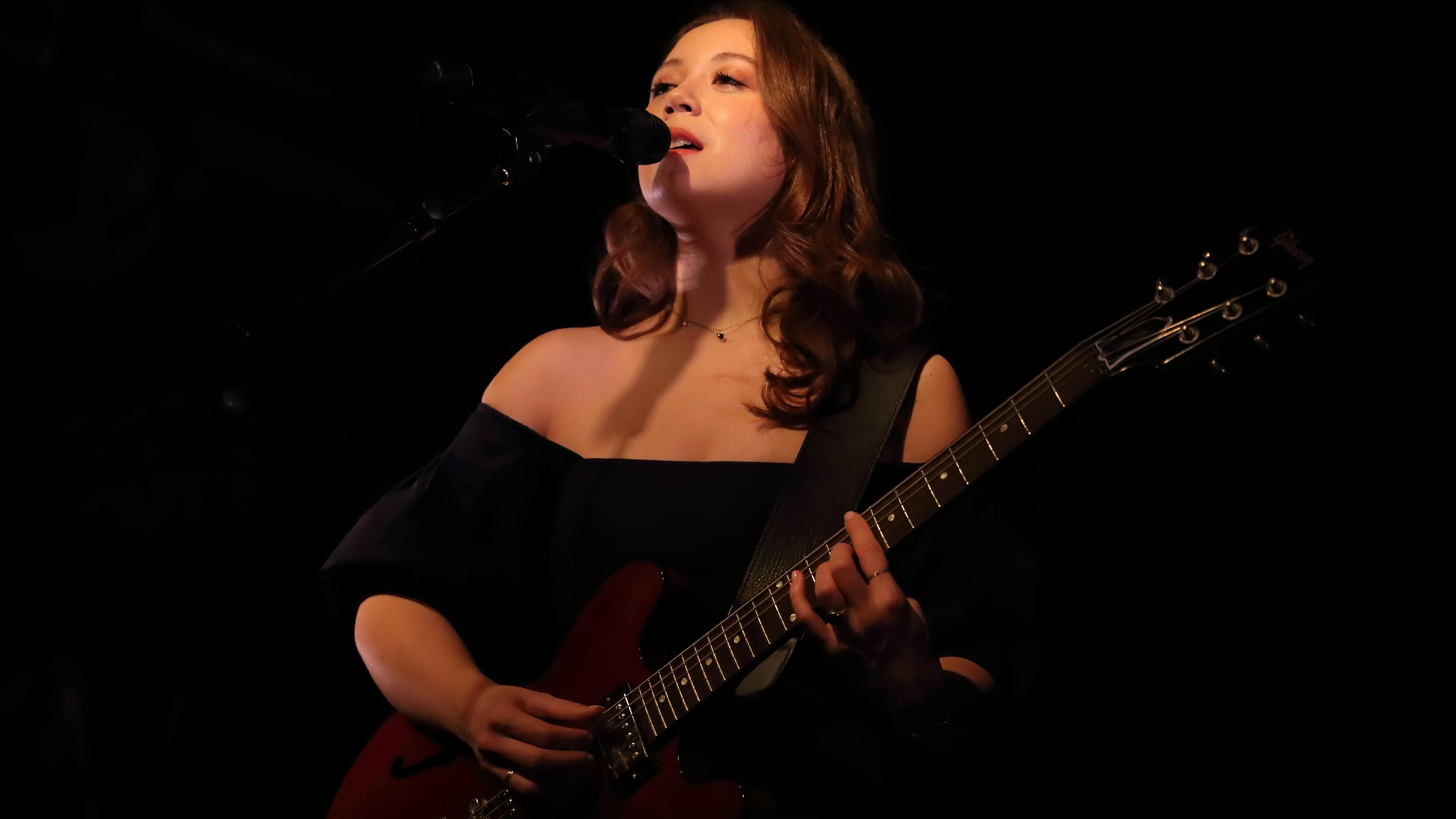 Eine Frau steht auf einer Bühne, spielt Gitarre und singt dazu.