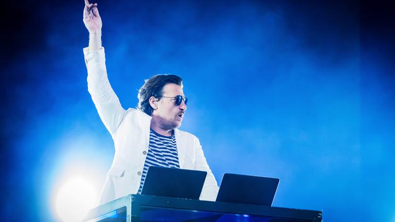 Ein Mann, der einen weißen Anzug trägt, streckt den Arm empor. Er steht hinter einem DJ-Pult auf einer Bühne.