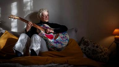 Eine blonde Frau sitzt in einem abgedunkelten Raum auf einem Bett und spielt Gitarre.