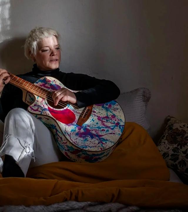 Eine blonde Frau sitzt in einem abgedunkelten Raum auf einem Bett und spielt Gitarre.